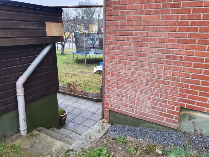 Fuktfläckar på grunden och tegelfasaden vid husknut, med stuprör och bakgård i bakgrunden.