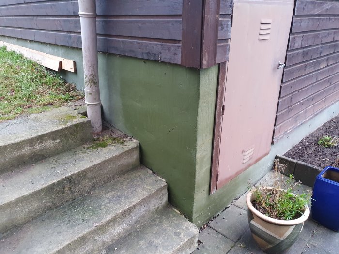 Fuktfläckar på grönmålad grund och tegelfasad vid husknut med avloppsrör och trappa.