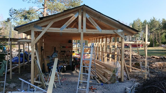 Pågående bygge av en träkonstruktion liknande ett garage med öppna väggar och synligt virke, under en klarblå himmel.