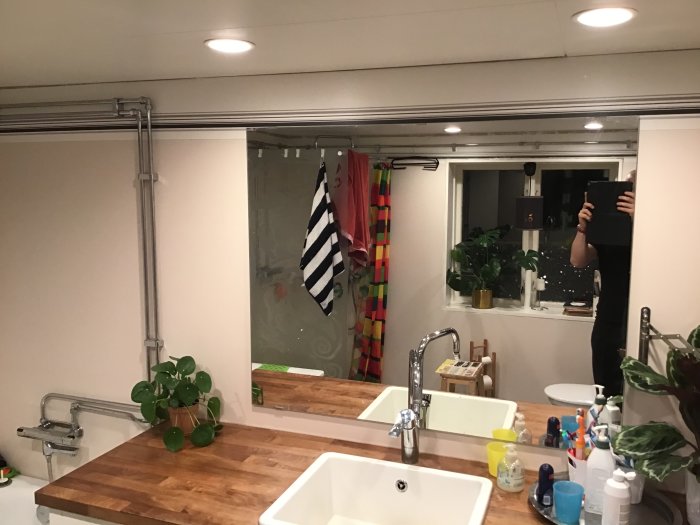 Badrum interiör med spegel och handfat, väggen öppen bredvid, spotlights i taket, person speglar sig med surfplatta.