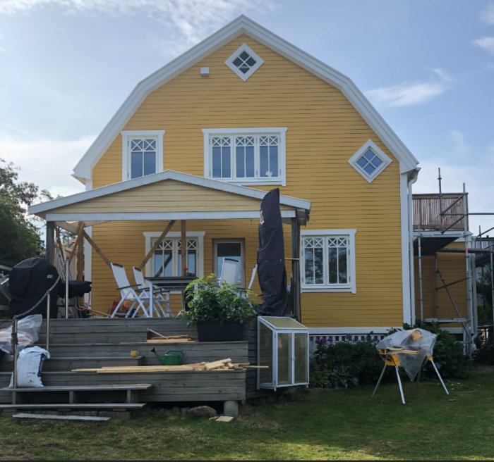 Gul villa med befintlig terrass och trädäck under renovering, omgivet av byggmaterial och verktyg.