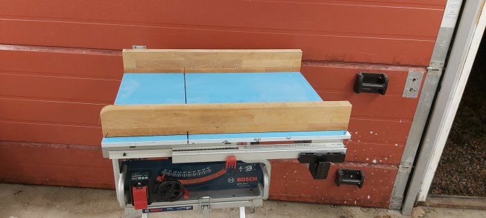 Blå Bosch bordssåg GTS 10 J med originalstativ framför en röd garagedörr, repor synliga på sågbordet.