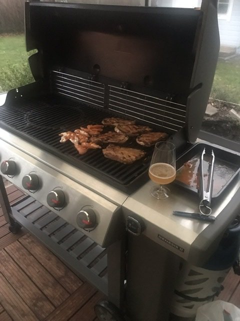 Weber Genesis E-610 grill med kött på grillytan, ölglas och grilltillbehör bredvid i trädgårdsmiljö.