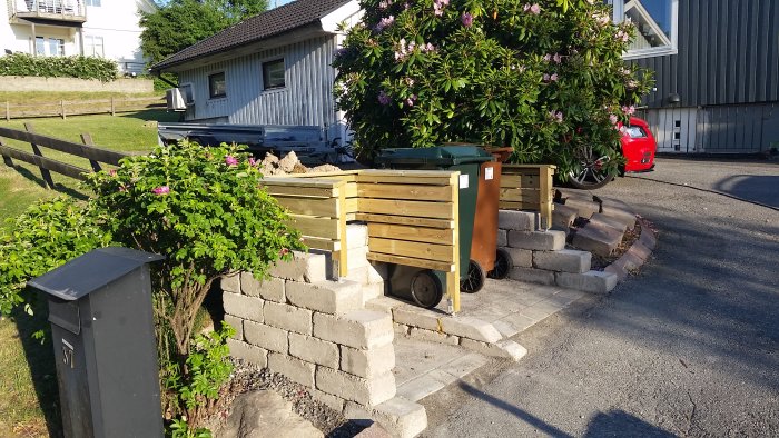 Trädgård med stentrappa och virkesklädd soptunneskydd.