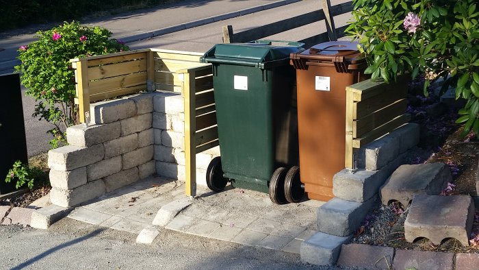 Ett DIY sopkärlsförråd gjort av stenblock och träplankor, med tre sopkärl inuti och växter i bakgrunden.