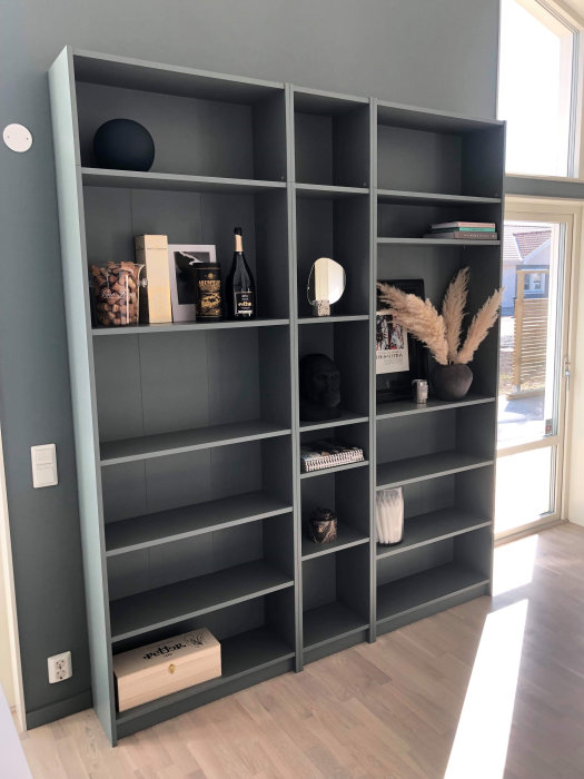 En stor bokhylla målad i grå/grön färg med diverse föremål i ett ljust rum.