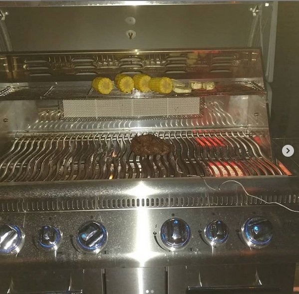 Grill med tända brännare och en köttbit på grillgallret samt grönsaker på övre hyllan.