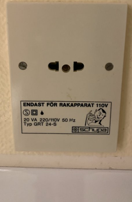 Gammalt rakapparatuttag på vägg märkt med "ENDAST FÖR RAKAPPARAT 110V", tillverkardetaljer och varningssymboler.