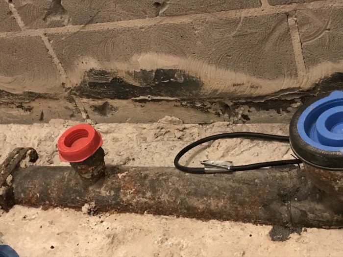 Gammalt korroderat gjutjärnsrör med rostfläckar och en röd rörändskap nära en betongvägg.