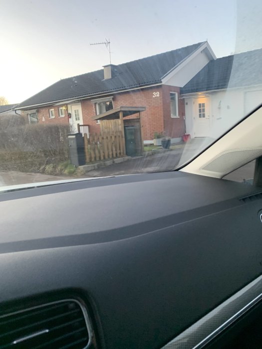Bild från bilfönster visar grannens tegelhus med ett sopkärlshus vid infarten.