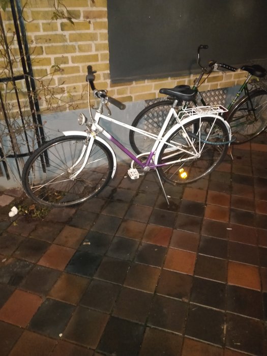 Vit lila damcykel och svart herrcykel parkerade på tegelstensgång vid en tegelvägg.