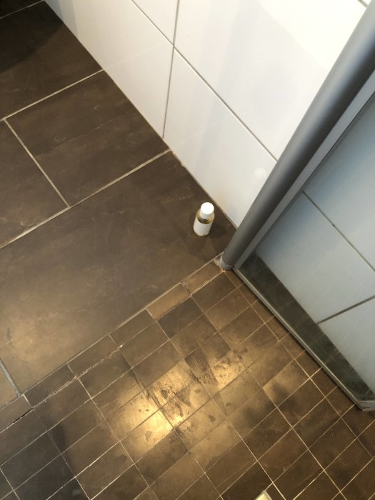 Brunaktiga kakelplattor på ett duschgolv med synliga slitage och sprickor i fogarna intill en duschvägg och flaska.