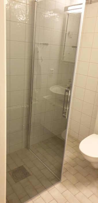Hörndusch med glasdörrar och vita kakelväggar i ett badrum med synlig toalett.
