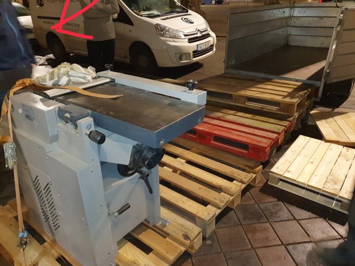 Maskin på pallar framför en lastbil, person med händer i fickorna i bakgrunden.