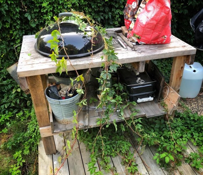 Klotgrill inbyggd i ett hemmagjort träbord omgivet av grönska, med grilltillbehör och en gasflaska intill.