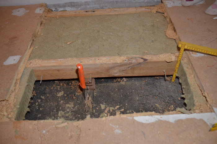 Öppen golvsektion med synlig betong, träreglar och isolering, mätverktyg syns.