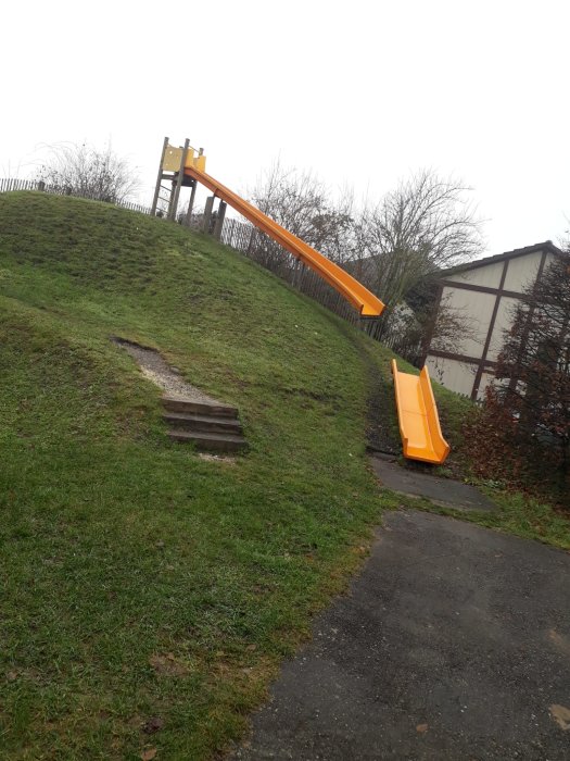 Orange rutschkana på en gräsbevuxen kulle med trappor, molnig himmel i bakgrunden.