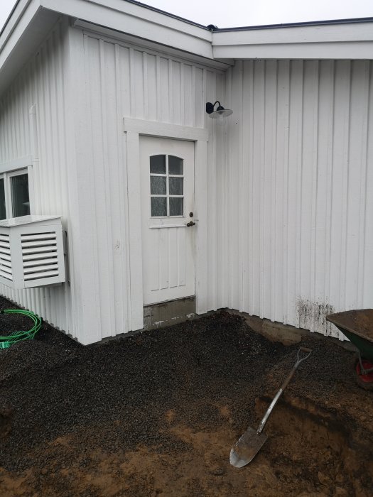 Utgrävning för utbyggnad intill hus med vit fasad, en dörr och utspilld makadam nära grunden.