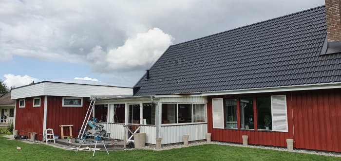 Ett hus med rött träpanel och svart takbeklädnad med pågående renoveringsarbete, inklusive stege och såg.