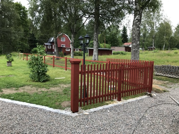 Nybyggt rött trästaket och grind framför ett hus i lantlig miljö, kantsten och grusgång syns i förgrunden.