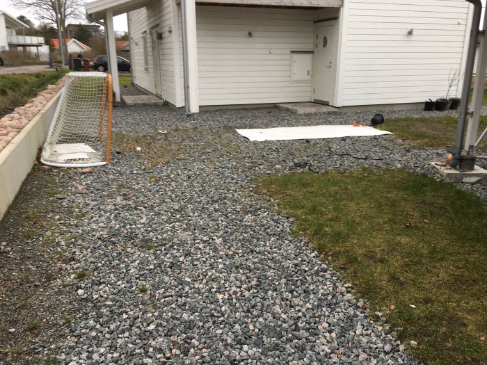 Område med makadam bredvid en carport intill en gräsplätt, planerat för marksten och rabattbyggnation.