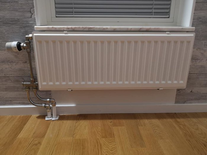 Nyinstallerad vit radiator under ett fönster med synliga kopplingar på sidan och rör som löper längs golvet.