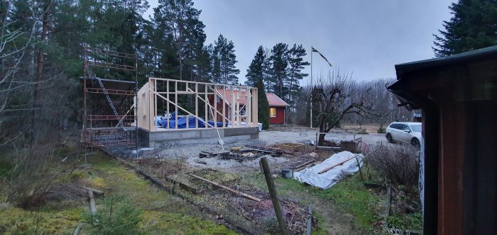 Nybyggd trästomme för hus på byggplats med byggställning och svensk flagga, omgiven av träd och befintligt hus.