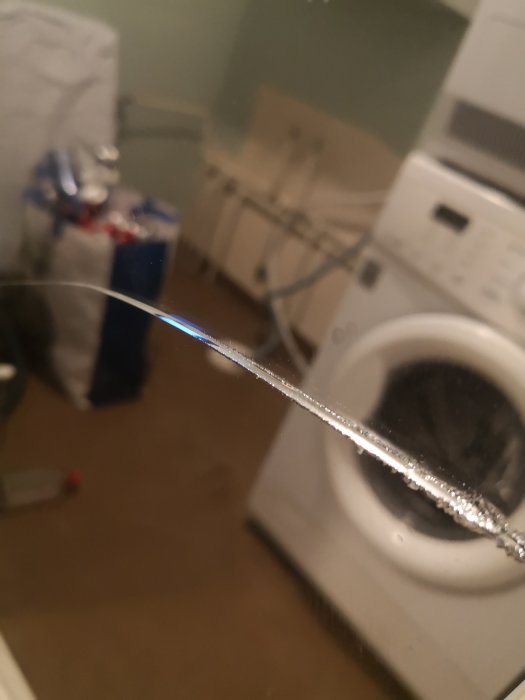 Spegel med en tydlig spricka i närbild med oklar bakgrund av en tvättmaskin och tvättstuga.
