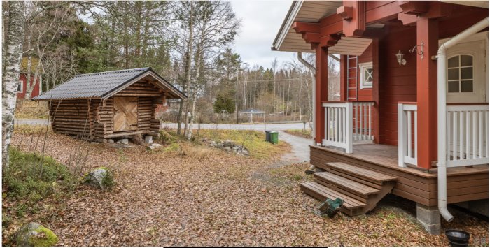 Rödmålat hus med veranda och lampa vid dörren, vedbod till vänster, höstlöv på marken.