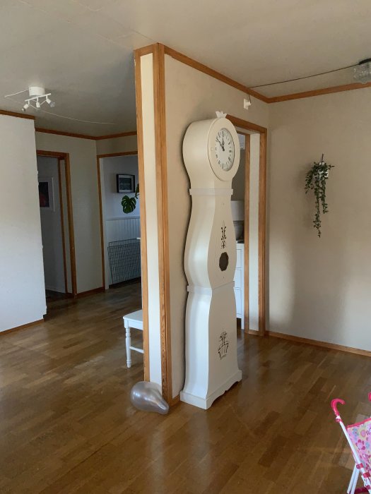 Del av ett vardagsrum i ett modulhus med en stor vit golvklocka och en synlig väggsektion och takbjälke som kan vara bärande.