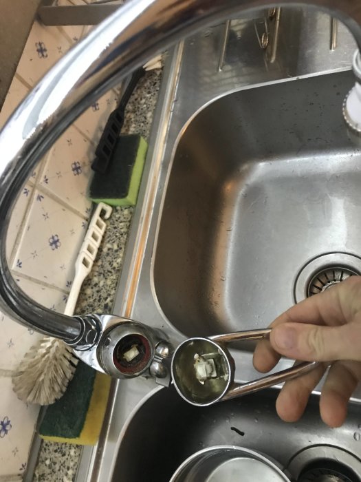 Närbild av trasig köksblandare som hålls isär, med fokus på slitna inre komponenter och bakgrundens diskho.