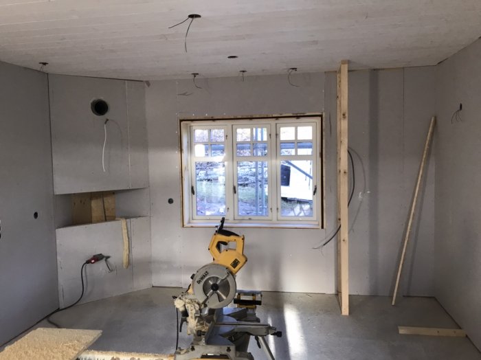 Byggarbetsplats inomhus med omonterat kök, synliga gipsskivor och skärmaskin framför fönster.