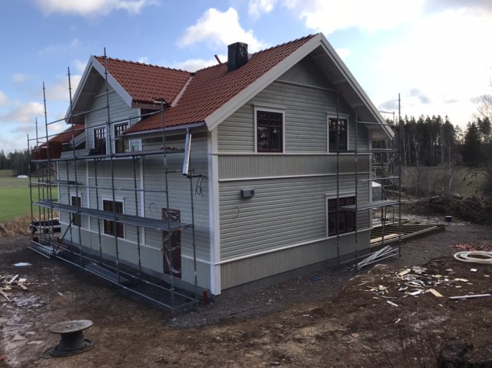 Nybyggd tvåvåningsvilla med byggställningar och nästan färdigställd fasad omgiven av ett oanlagt trädgårdsområde.