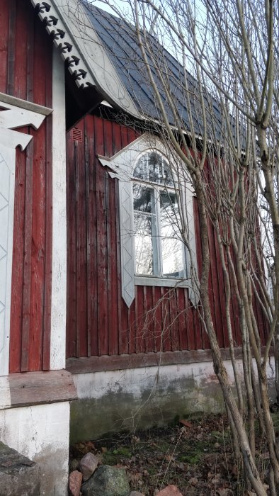 Rödfärgad byggnad med dekorativa snickeridetaljer och stort originalfönster, på krypgrund omgiven av naturen.