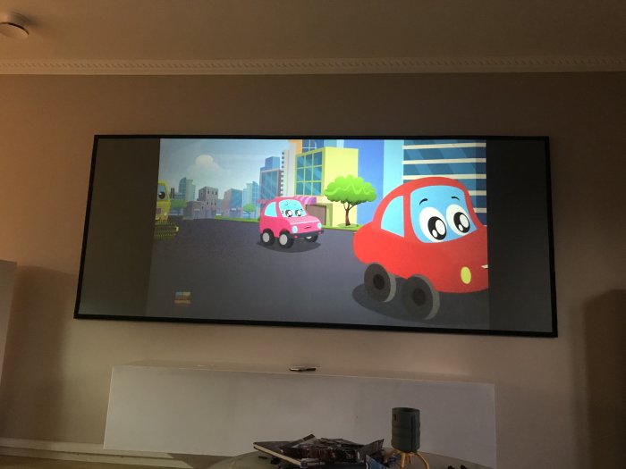 Ramspänd projektorduk på vägg med animerat bilprogram, ovanför en vit möbel i ett vardagsrum.