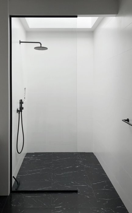 Modern dusch med vit kakel, svart golv och ett fönster högst upp som släpper in ljus.