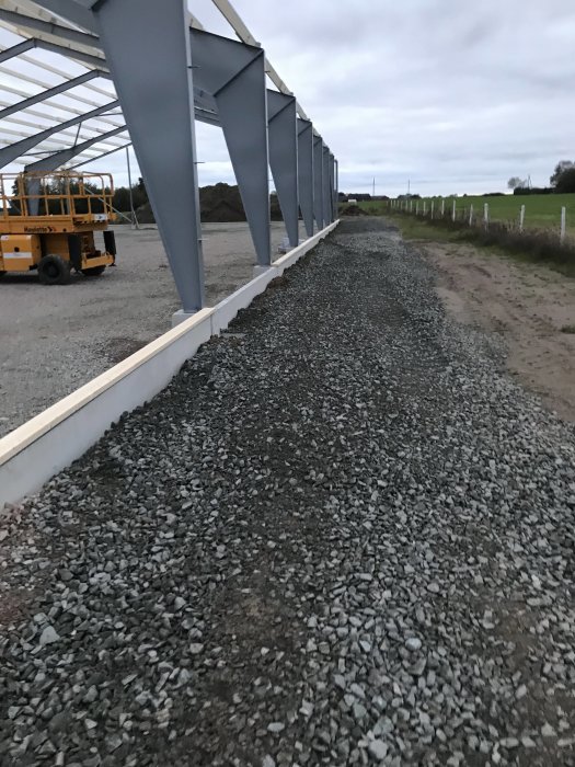 Stålstomme för en byggnad under konstruktion, gruslagd mark vid kantbalken och en terränggående lift parkerad på sidan.