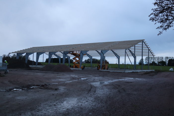 Stålstomme under uppbyggnad med lyftkran och arbetslyft på en lerig byggplats, omgiven av högar med material.