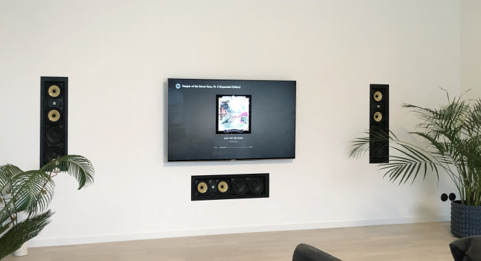 Vardagsrum med väggmonterad TV och inbyggda högtalare, inredning inkluderar krukväxt.