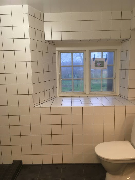 Nyrenoverat badrum med vita kakelväggar, ett fönster och en toalettstol, i väntan på städning och inredning.