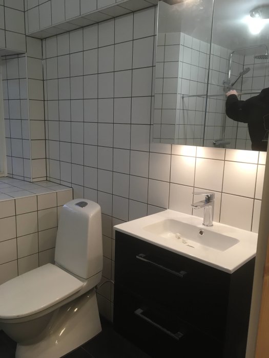 Renoverat badrum med vit kakling, WC och svart kommod med handfat och spegelskåp.