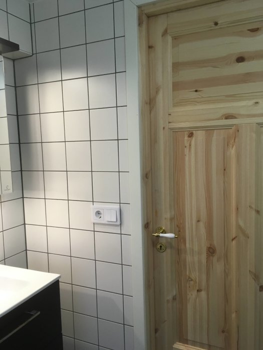 Hörn av nyrenoverat badrum med vita kakelväggar, en omålad träddörr och svart skåp.