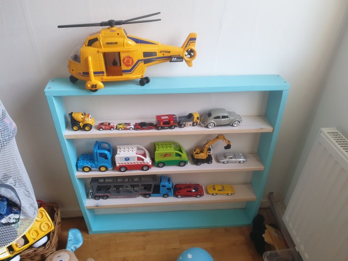 Hemmagjord hylla i ljus MDF med leksaksbilar och helikopter, inomhus bredvid en radiator.