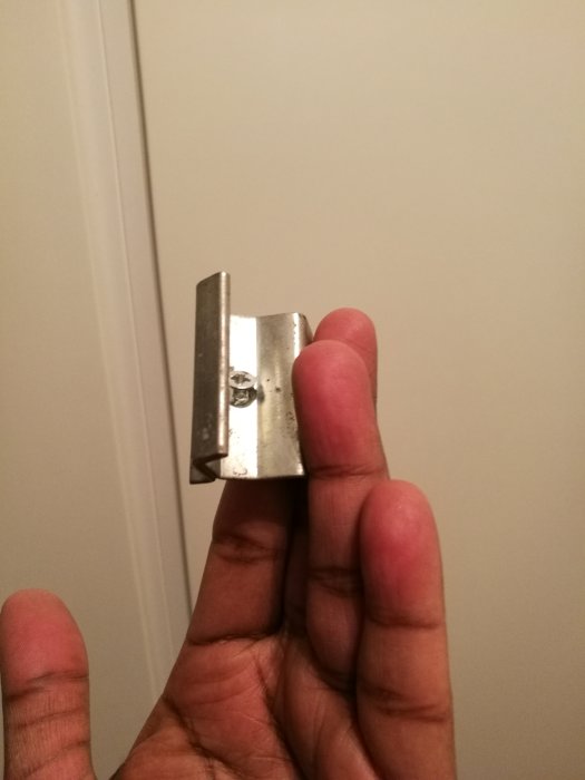 Hand håller ett metallbeslag med pegg för vädringslucka, mot vit dörrbakgrund.