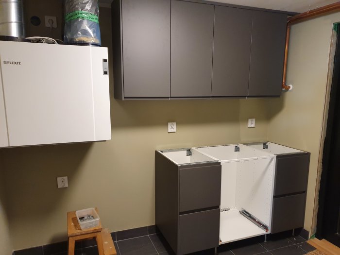 Ett ofärdigt kök med installerade underskåp och väggskåp, ett FTX-ventilationsaggregat och avklippta rör.