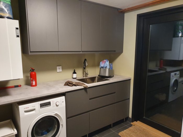 Kök med gråa IKEA-skåp, diskbänk och vitvaror inklusive tvättmaskin, med svart dörrkarm.