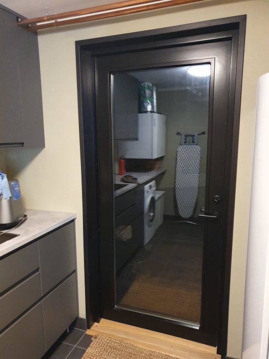 Utsikt genom svart dörröppning till ett tvättstuga med IKEA-skåp, tvättmaskin och värmepumpsinstallation.