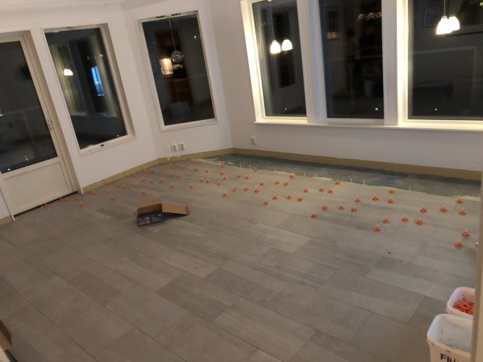 Pågående läggning av grått klinkergolv med oranga distanskilar och en kartong på golvet.