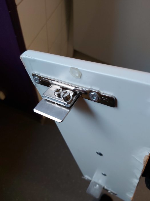 Närbild av en vit toalettstol med ett öppet metallfäste och synliga skruvhål där en del saknas.