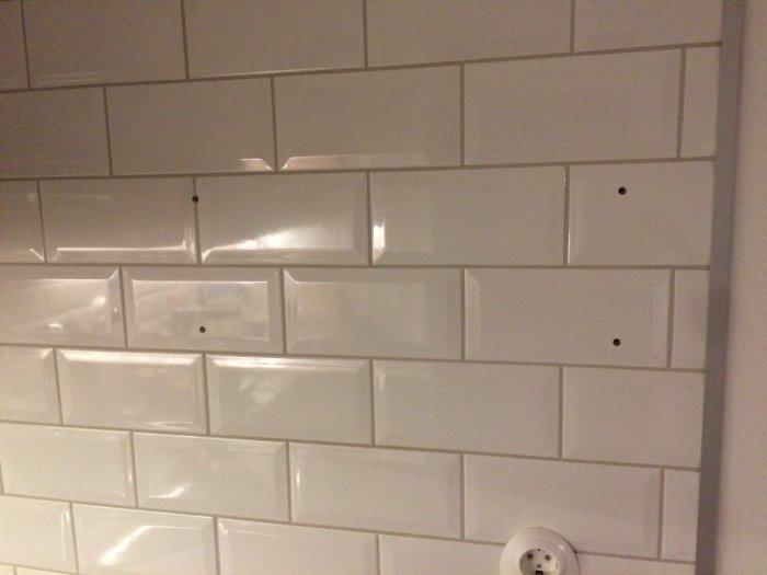 Vägg med vita kakelplattor och fyra prydliga borrmärken för hyllmontering.